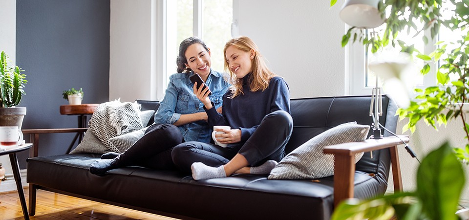 Happy young female friends sitting together on couch in living with a mobile phone. Two young women relaxing at home using mobile phone.