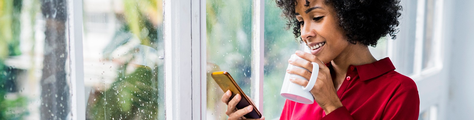 Woman drinking coffee and looking at cellphone while it rains outside