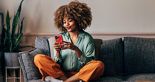 A smiling elegant African-American female using her smartphone while sitting on the cozy sofa in the living room.