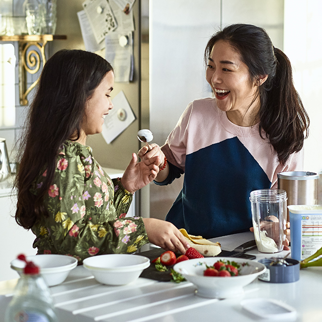 Woman holding yoghurt on spoon towards girl, laughing, threatening to smear food on daughter's face, messing about, having fun, playing, bonding